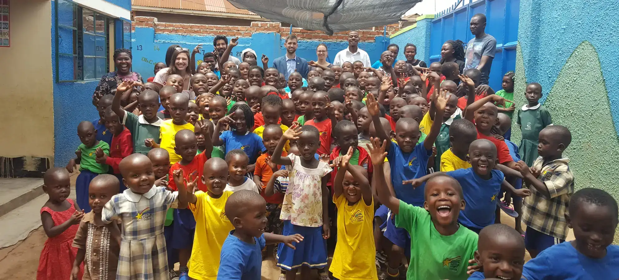 Dozens of Little Light children in colourful shirts waving in the schoolyard in Namuwongo, Kampala, with volunteers and staff behind them